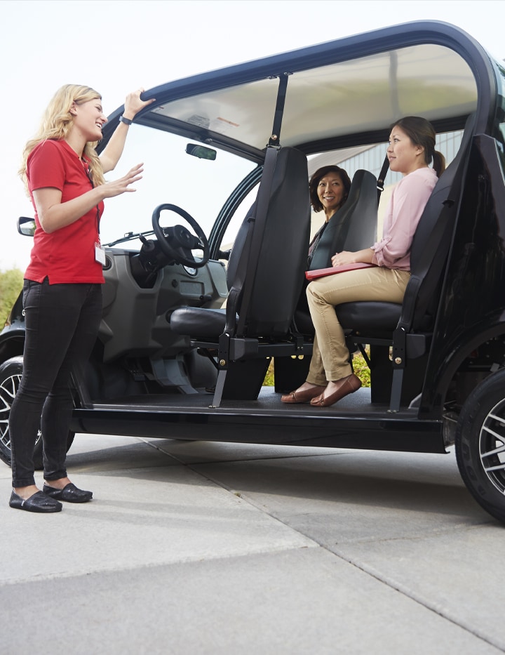 A woman giving standing next to an electric vehicle giving direction to two passengers