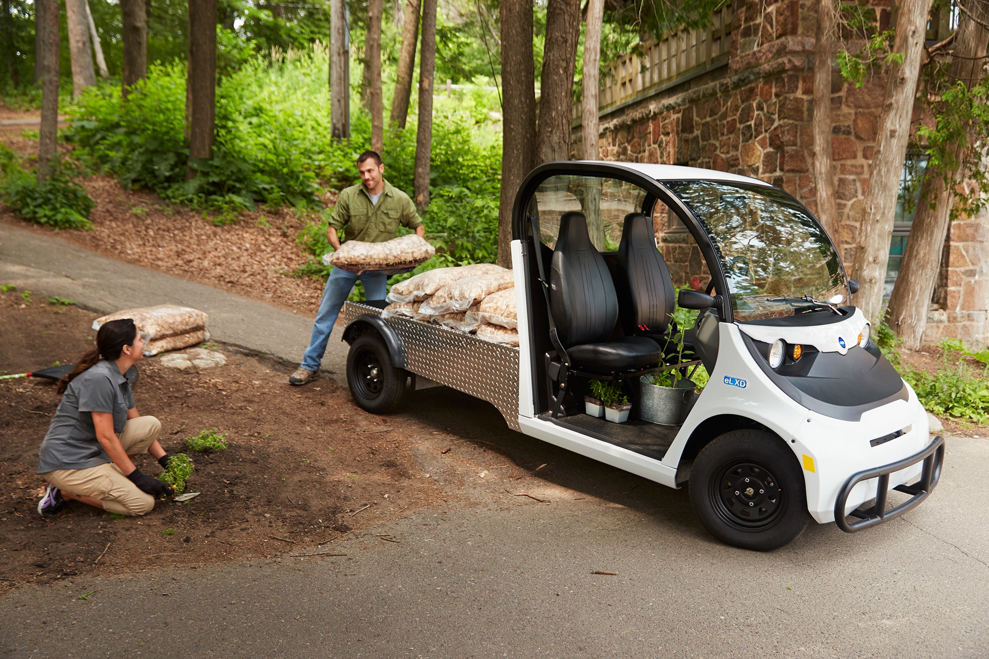A man unloading bags of mulch from an electric vehicle next to a woman planting in the park