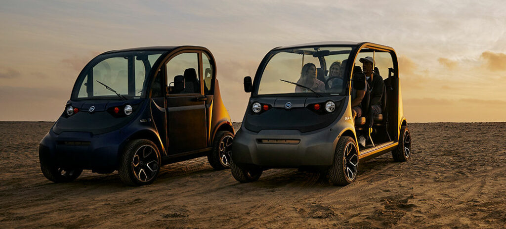 Two side by side GEM electric vehicles on the beach, one full of people and one empty